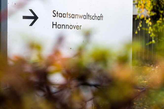 Ein Hinweisschild mit der Aufschrift "Staatsanwaltschaft Hannover" steht hinter einer Hecke, deren Herbstfarben verschwommen im Bildervordergrund erkennbar sind.