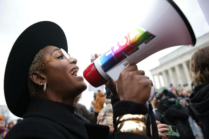 Weiblich, schwarz gelesene Person mit schwarzem Mantel, Brille und breitkrempigen Hut spricht in ein mit regenbogenfarben Folien verzierten Megafon, im Hintergrund ist eine Demonstration und das Lincoln Memorial zu sehen.