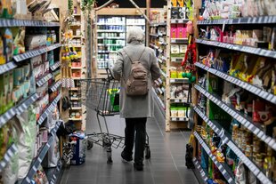 Eine Frau geht uns den Rücken zugewand mit ihrem Einkaufswagen durch eine Reihe Supermarktregale