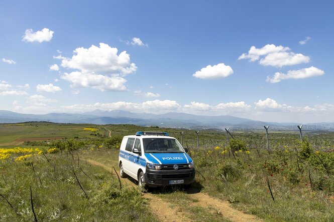 Ein deutsches Polizeiauto patrolliert an einem Grenzzaun in einer grünen Landschaft