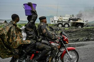 Drei Männer fahren auf einem Motorrad an einem brennenden Panzerfahrzeug vorbei