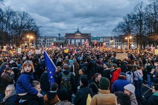 Eine große Menschenmenge steht in der Abenddämmerung vor dem Brandenburger Tor, viele halten Plakate, Fahnen und kleine Lichter in die Höhe