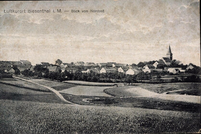 Ein historisches Schwarz-Weiß-Foto zeigt ein Dorf mit Kirche, davor Felder