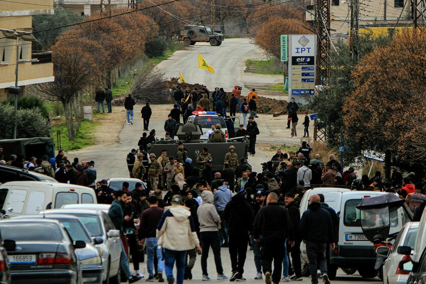 Menschen stehen hinter gepanzerten Fahrzeugen der libanesischen Armee, während eine Sandbarriere und ein israelischer Militärjeep im Hintergrund die Straße am Rande des südlibanesischen Dorfes Kfar Kila blockieren