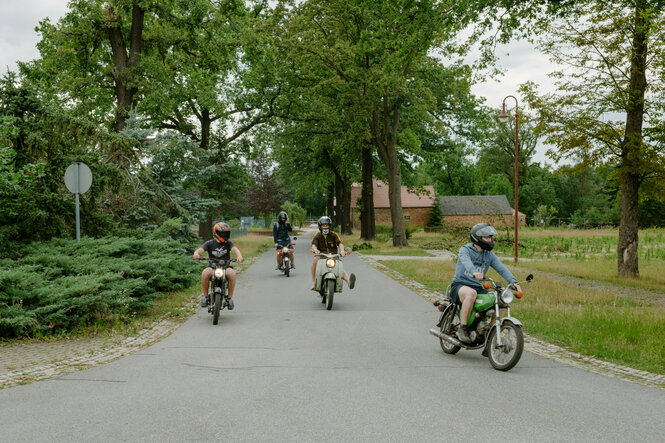 Mehrere Personen fahren auf Mopeds eine Dorfstraße entlang.