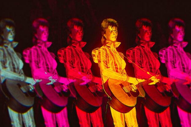 Filmstill: Eine Aufnahme von David Bowie bei einem Konzert ist multipliziert und abwechselnd in Grün- Pink-, Rot- und Orangetönen getaucht. Er spielt Gitarre und trägt ein schimmerndes Outfit mit Stehkragen.