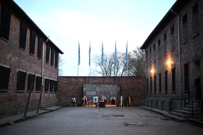 Sieben Blumenkränze stehen an einer Wand, vor der viele Kerzen brennen, in Erinnerung an die ermordeten Häftlinge