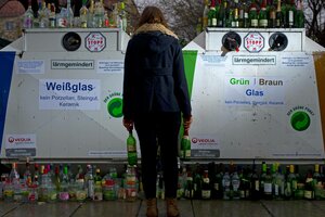 Eine Person steht mit Leergut vor einem vollen Glascontainer.