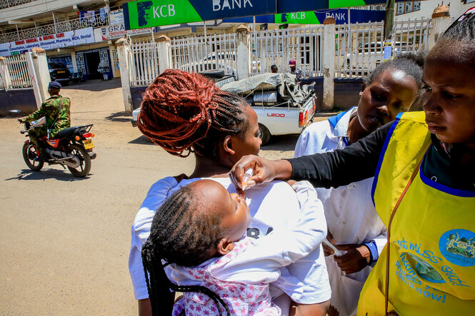 Mutter mit Kind bei Impfung, mitten auf einer staubigen Straße in einem Slum in Nairobi. Dem Kind, das auf dem Rücken seiner Mutter ist, wird das Serum von zwei Gesundheitsarbeiterinnen, in den Mund geträufelt