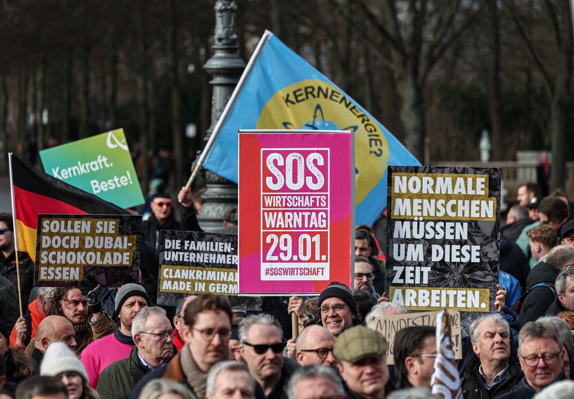 Gegendemonstranten halten dunkle Schilder mit Aufschrift «Normale Menschen müssen um diese Zeit arbeiten» und «Sollen sie doch Dubai-Schokolade essen» auf der Kundgebung «Wirtschaftswarntag» vor dem Brandenburger Tor hoch.