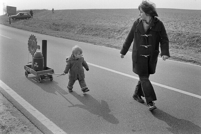 Ein Kind zieht ein Wägelchen mit einem Modell AKW über eine Asphaltstrasse, die Mutter dreht sich zum Kind