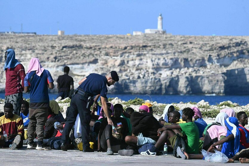 Migranten sitzen in der prallen Sonne auf der Straße und warten. Vor ihnen steht ein Mann in Uniform, der sich zu einem der Migranten beugt