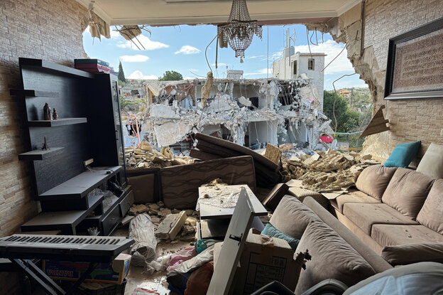 Blick aus einem Wohnzimmer, in dem es keine Fenster sondern ein Loch gibt, auf ein zerstörtes Gebäude gegenüber. In der Wohnung stehen Sofas, eine Regal, ein Keyboard, alles liegt durcheinander und ist verstaubt. An der Decke hängt noch der Kronleuchter