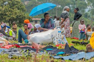 Eine Gruppe von Frauen sitzt uns steht mit ihren Kindern in der freien Natur, es regnet, eine Frau stützt sich auf einen Sack und schützt sich mit einem Regenschirm