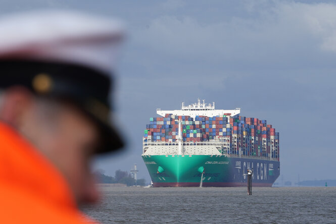 Ein Containerschiff befährt die Elbe, im Vordergrund ein Mann mit Signalweste