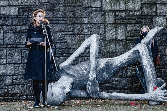 Frau spricht ins Mikrofon neben der Skulptur eines abgemagerten Menschen