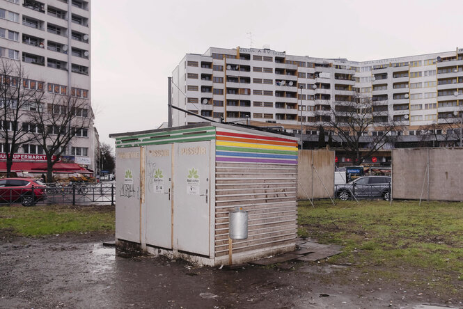 die Ökotoilette am Kottbuser Tor in Berlin-Kreuzberg von außen: ein weißer Holzbau mit aufgemalter Regenbogenfahne