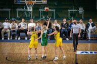 Drei Basketballerinnen bei einer Szene vor dem Basketballkorb, im Hintergrund eine mit Männern besetzte Zuschauerbank