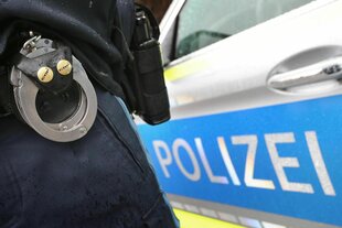 Links im Bild der eine Polizistenbein mit Handschellen, rechts davon ein Polizeiauto