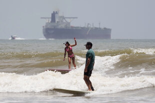 Ein Öltanker verlässt den Hafen; im Vordergrund surfen zwei Menschen