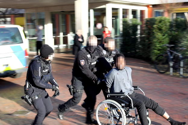 Polizeibemate schieben einen Schwarzen Mann in einem Rolltuhl von links nach rechts durchs Bild. Im Hintergrund ist ein Krankenhaus zu erkennen