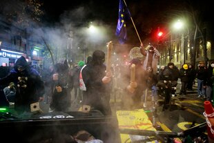 Menschen, zum Teil maskiert, auf einer nächtlichen Straße hinter einer Barrikade. Einer hält eine EU-Fahne in die Höhe