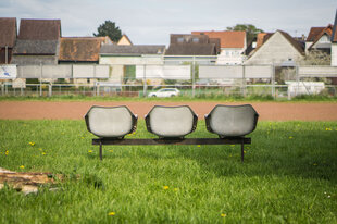 Verlassen stehen neben einem Dorfsportplatz drei Sitzplätze am Spielfeldrand.