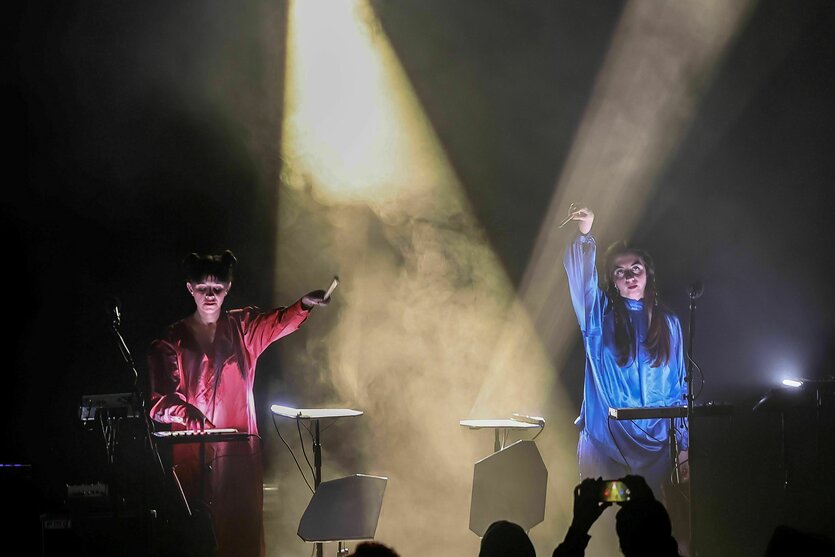 Zwei Frauen in roter und blauer Kleidung stehen an Musikinstrumenten auf einer von Nebeln umwobenen Bühne