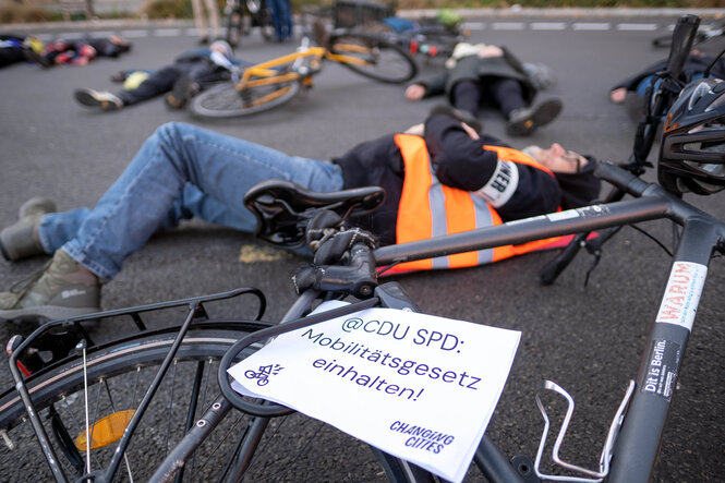 Menschen liegen bei einem Protest auf der Straße. Auf einem Plakat steht: "@CDU SPD: Mobilitätsgesetz einhalten!"