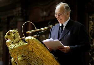 Der Aga Khan steht lächelnd an einem Rednerpult, das mit einer Adlerskulptur mit gebreiteten Schwingen geziert ist