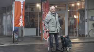 Eine Person steht mit einem Rollkoffer auf einer Straße, im Hintergrund der Eingang zu einem Laden. Vor dem Laden weht eine orangfarbene Fahne mit der Aufschrift 