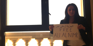 Linken-Abgeordnete Sevim Dağdelen steht in Saudi-Arabien vor einer Fensterbalustrade und demonstriert mit einem Schild in der Hand, auf dem steht "Free Ali Al Nimr".