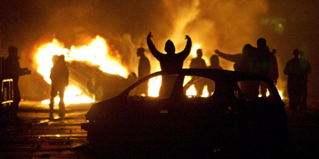 Nächtliche Szene mit brennenden Autos und Protestierenden.