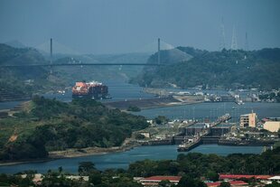 Ein Frachtschiff fährt durch den Panamakanal, im Vordergrund ist eine Schiffschleuse, im Hintergrund eine Hängebrücke und sonst nur tropische Grüne.