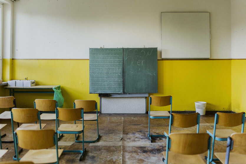 Stühle stehen in einem Klassenraum mit Tafel, der Boden ist wellig und alles macht einen leicht ramponierten Eindruck