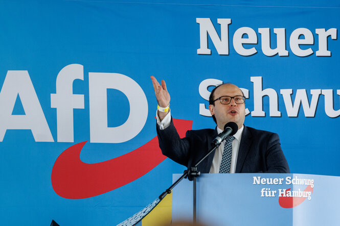 Krzysztof Walczak spricht auf einer Bühne vor einem AfD-Plakat