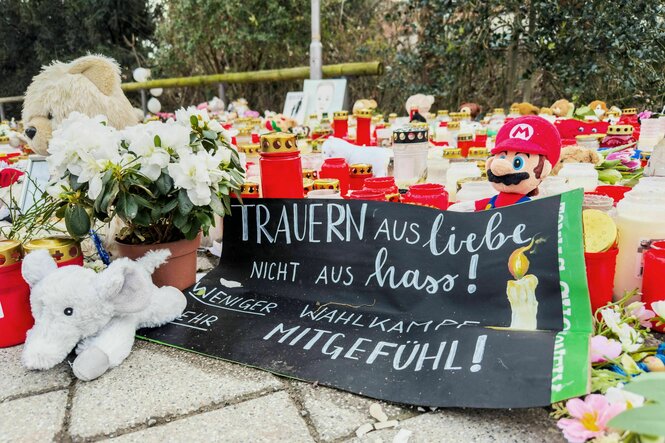 Zahlreiche Blumen, Kuscheltiere und Kerzen liegen am Tator in einem Park in Aschaffenburg. Dazwischen ein Plakat, auf dem steht: "Weniger Wahlkampf, mehr Mitgefühl"