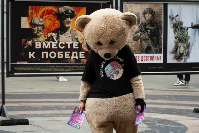 Eine Person in traurig wirkendem Bärchenkostüm vor Plakaten mit russischer Kriegspropganda