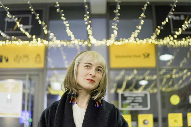 Foto der Yelizaveta Landenberger vor schillerndem Deutschen Post Hintergrund. Sie hat ein freundliches Gesicht, vorne anblondiertes Pony und verspielte bunte Ohrringe.