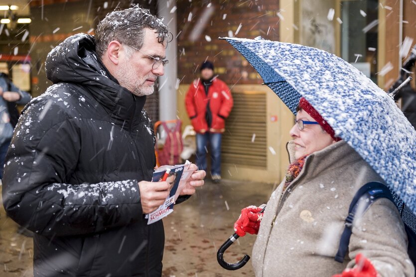 Wolfgang Schmidt steht im Schneefall einer Frau gegenüber, die sich mit einem regenschirm schützt