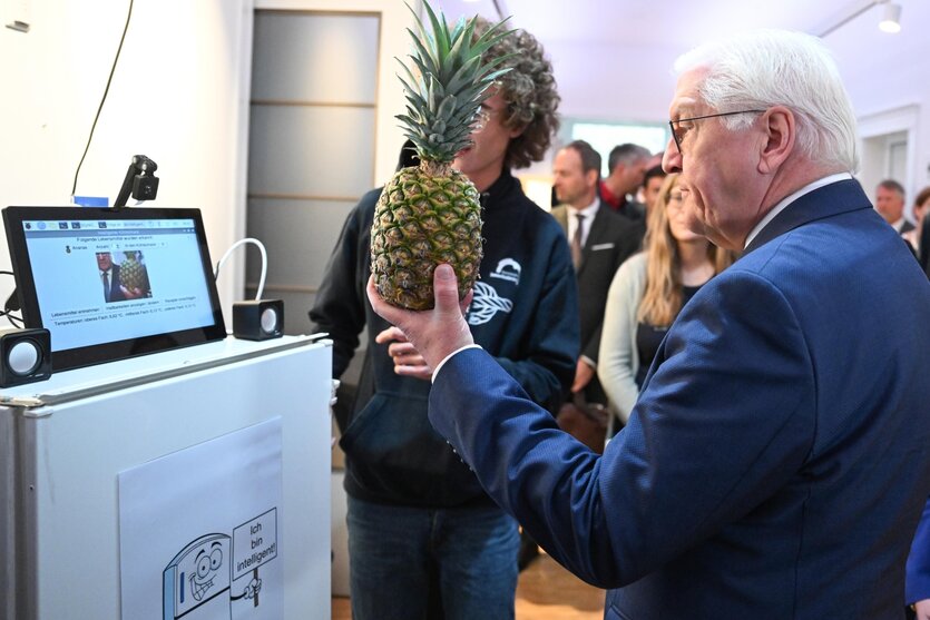 Mann hält, umringt von weiteren Menschen, eine Ananas vor einen Bildschirm