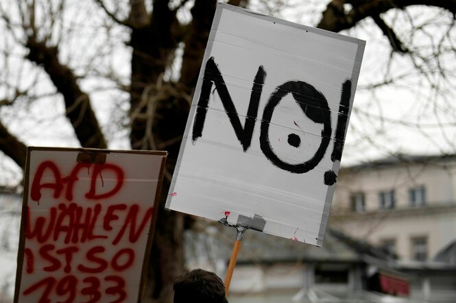 Zu sehen sind zwei Protestschilder. Auf einem steht "AfD wählen ist so 1933", das andere zeigt das englische Wort "No", wobei das O mit Frisur und Hitlerbärtchen an Adolf Hitler erinnern soll.