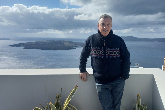 Ein Mann mit blauer Strickjacke steht auf einem Balkon. Im Hintergrund das Meer und eine Insel