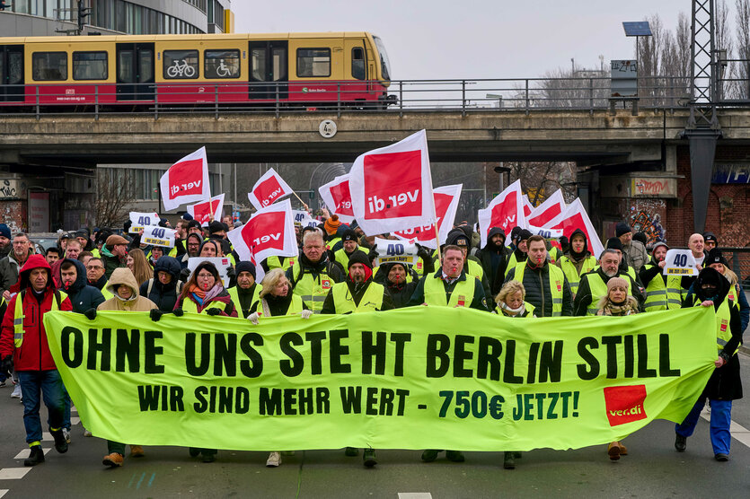 Demonstration mit Transparent und Fahnen unter einer S-Bahnbrücke.