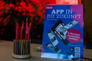 Nahaufnahme eines Aufstellers, der die taz App bewirbt. Er steht auf einem Tisch, daneben ein Stifthalter mit taz-Bleistiften.