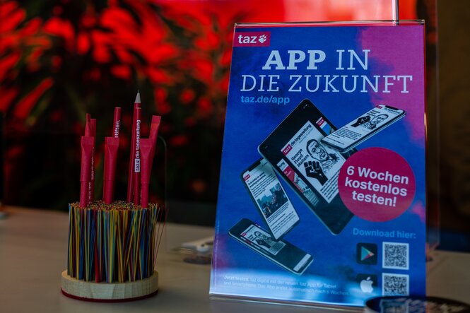 Nahaufnahme eines Aufstellers, der die taz App bewirbt. Er steht auf einem Tisch, daneben ein Stifthalter mit taz-Bleistiften.