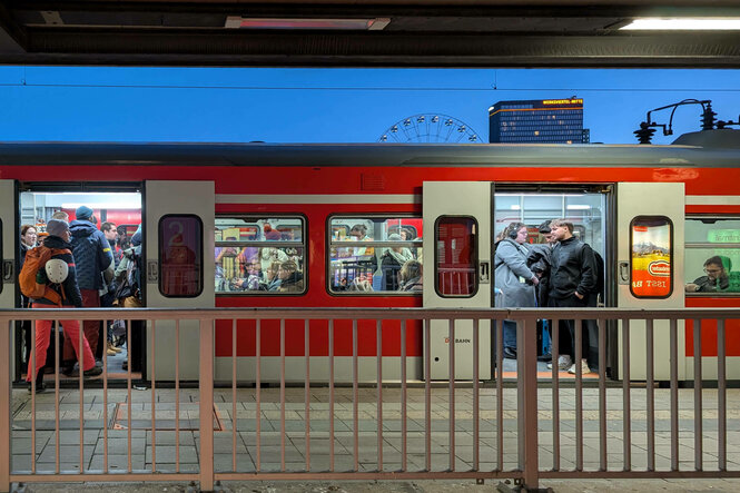 Eine S-Bahn steht an einem Bahnhof. Die Türen sind geöffnet. Drinnen einige Menschen, aber es ist nicht überfüllt. Im Hintergrund blauer Abendhimmel und ein Riesenrad.