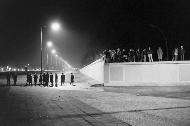 Menschen stehen auf der Berliner Mauer, Grenzbeamte stehen in einer Reihe im Mauerstreifen