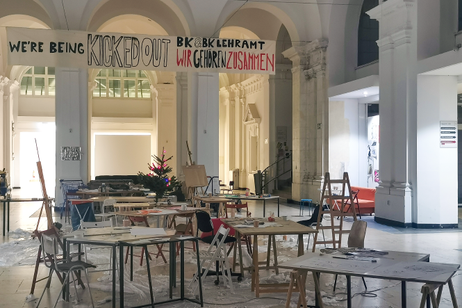 Ein chaotischer Raum in der UdK. An der Decke hängt ein Banner, auf dem steht: "We are being kicked out. BK & BK Lehramt Wir gehören zusammen"