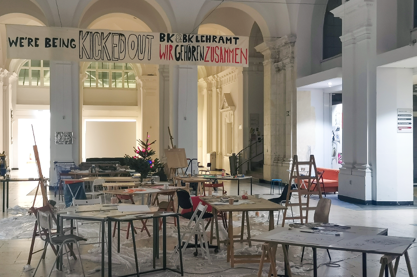 Ein chaotischer Raum in der UdK. An der Decke hängt ein Banner, auf dem steht: "We are being kicked out. BK & BK Lehramt Wir gehören zusammen"